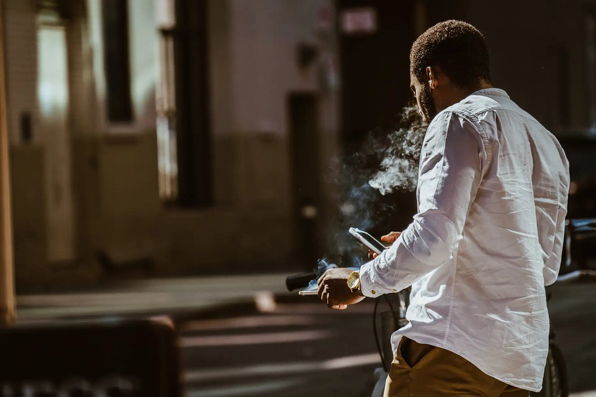 Mann im Freien, von hinten gesehen, hält ein Smartphone in der Hand, während er Rauch ausatmet; symbolische Darstellung eines COPD-Risikofaktors im Alltag.