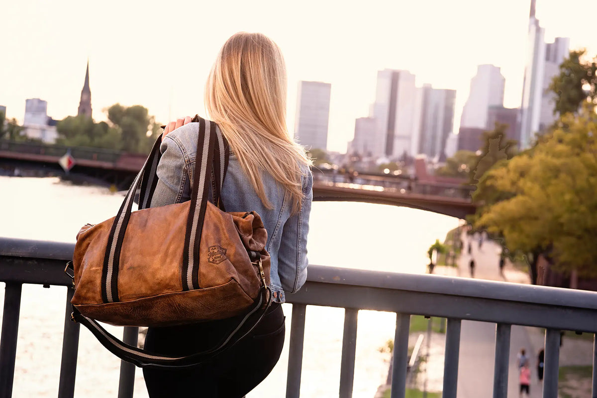 Frau steht auf einer Brücke mit dem Rücken zur Kamera und trägt einen ARTZT Vintage Series Weekender über der linken Schulter, während sie auf die Skyline von Frankfurt blickt