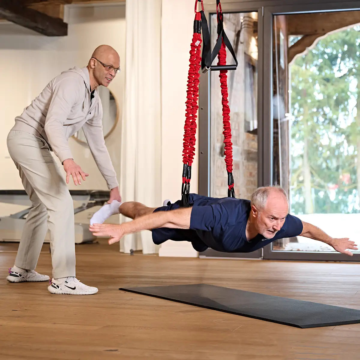 Therapeut begleitet eine Übung mit dem 4D PRO Bungee Schlingentrainer, während ein Mann mit dem Körper horizontal in den elastischen Seilen trainiert. #farbe_rot