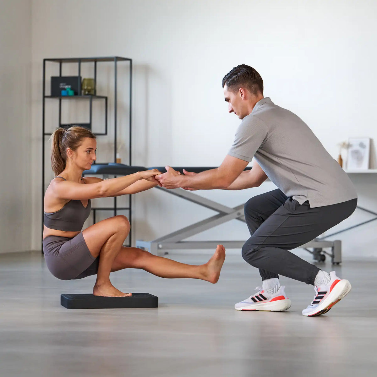 Frau führt eine einbeinige Kniebeuge auf dem Balance Pad von ARTZT thepro aus, während ein Trainer ihre Hände hält und die Stabilität im Physiotherapie- oder Trainingsraum unterstützt.