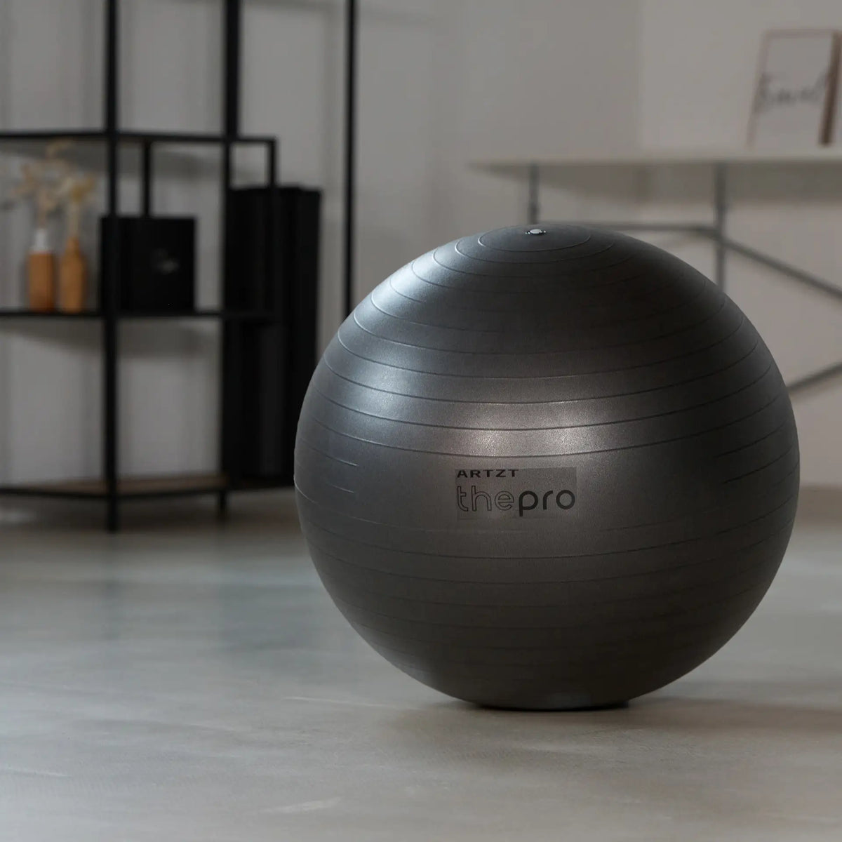Grauer Gymnastikball der Marke ARTZT thepro steht frei auf dem Boden eines Trainingsraums; die gerippte Oberfläche und das aufgedruckte Logo sind gut sichtbar und zeigen den Ball als vielseitiges Trainings- und Therapiegerät.