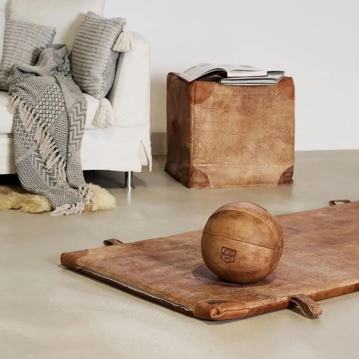 Ein ARTZT Vintage Series Medizinball liegt auf einer brauen Ledermatte, im Hintergrund stehen eine weiße Couch und ein brauner Sitzwürfel aus Leder.