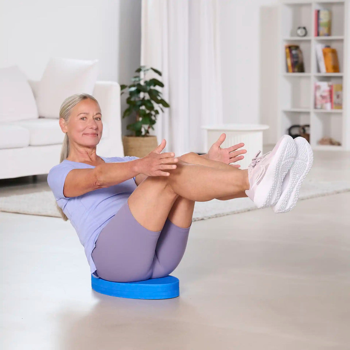 Frau trainiert im Wohnzimmer im Sitzen auf einem blauen Balance Pad von TheraBand