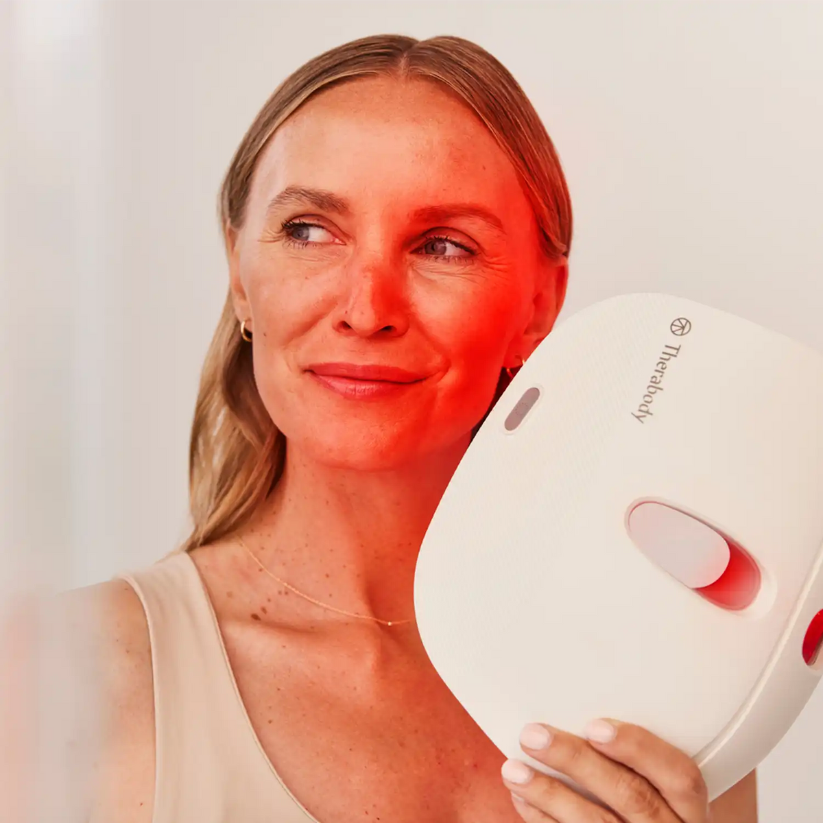 Frau blickt lächelnd zur Seite und trägt in der linken Hand eine Therabody TheraFace Mask LED-Maske.