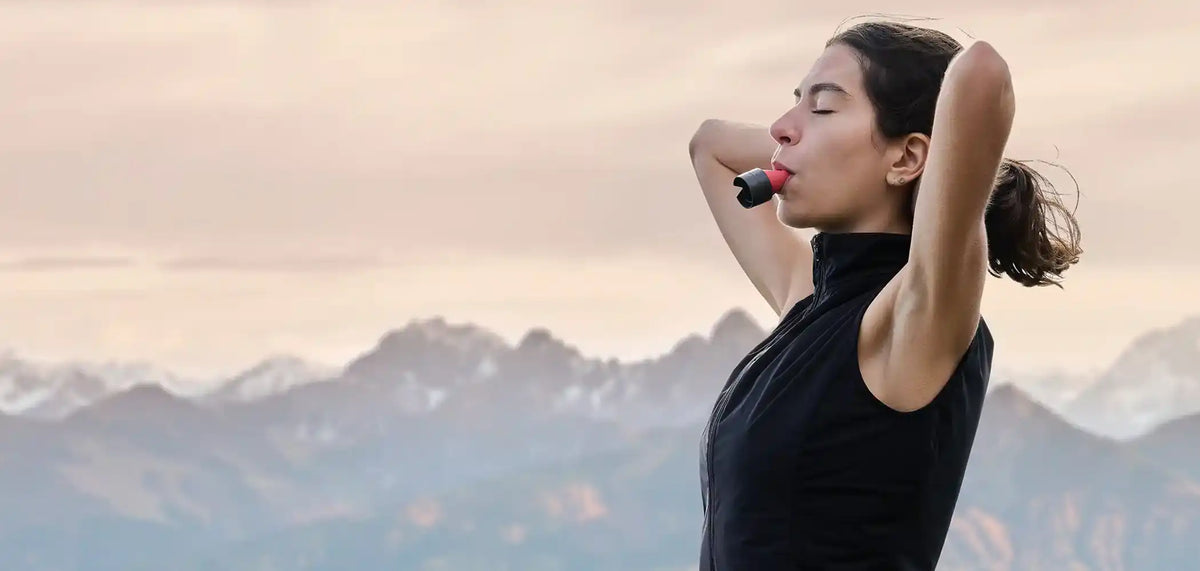 Eine Frau steht vor einem Bergpanorama und trägt den ARTZT neuro Atemtrainer im Mund, mit geschlossenen Augen und den Armen hinter dem Kopf verschränkt.