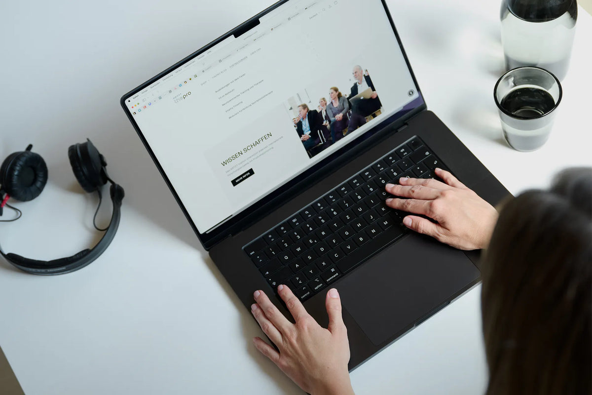 Ein geöffneter Laptop steht auf einer weißen Oberfläche. Links daneben liegt ein Paar schwarze Bügelkopfhörer, rechts daneben steht ein Glas und eine Glaskaraffe. Zwei Hände ruhen auf der Tastatur.