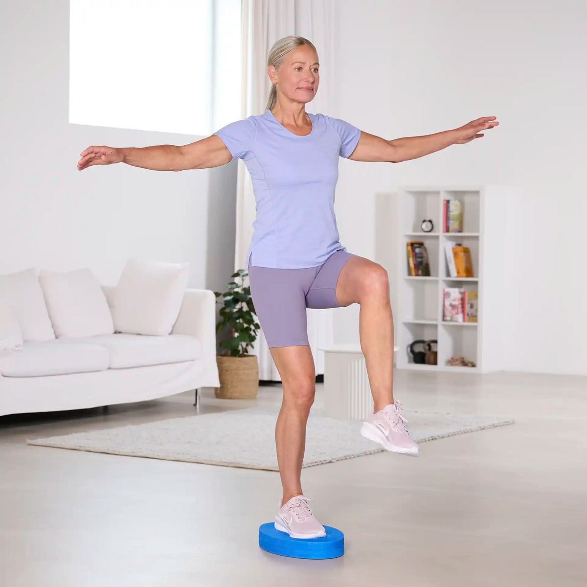 Frau im Wohnzimmer trainiert im Einbeinstand auf einem blauen Balance Pad der Marke TheraBand