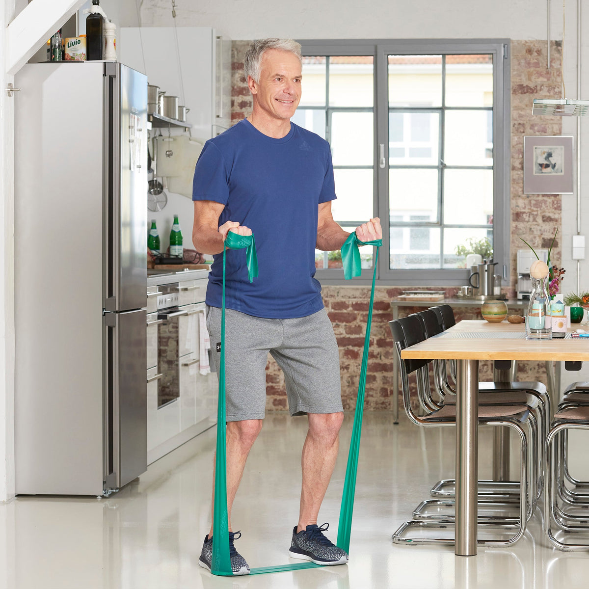 Sportler trainiert beide Oberarme im Stehen mit einem grünen TheraBand