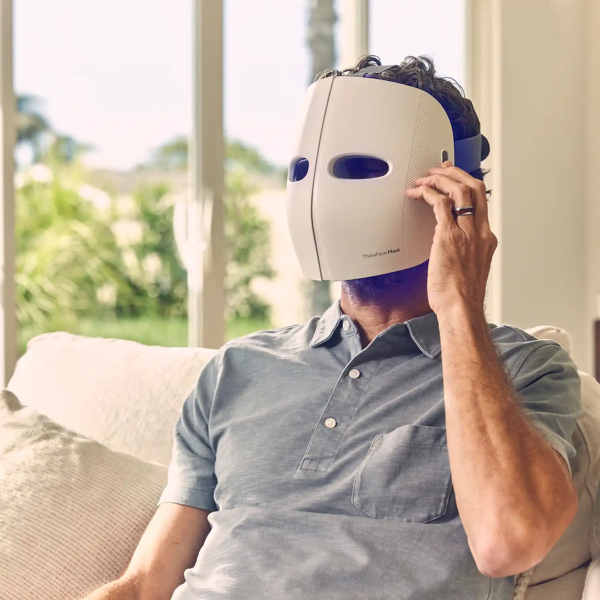 Mann sitzt auf einem Sofa und trägt eine Therabody TheraFace LED-Maske im Gesicht.